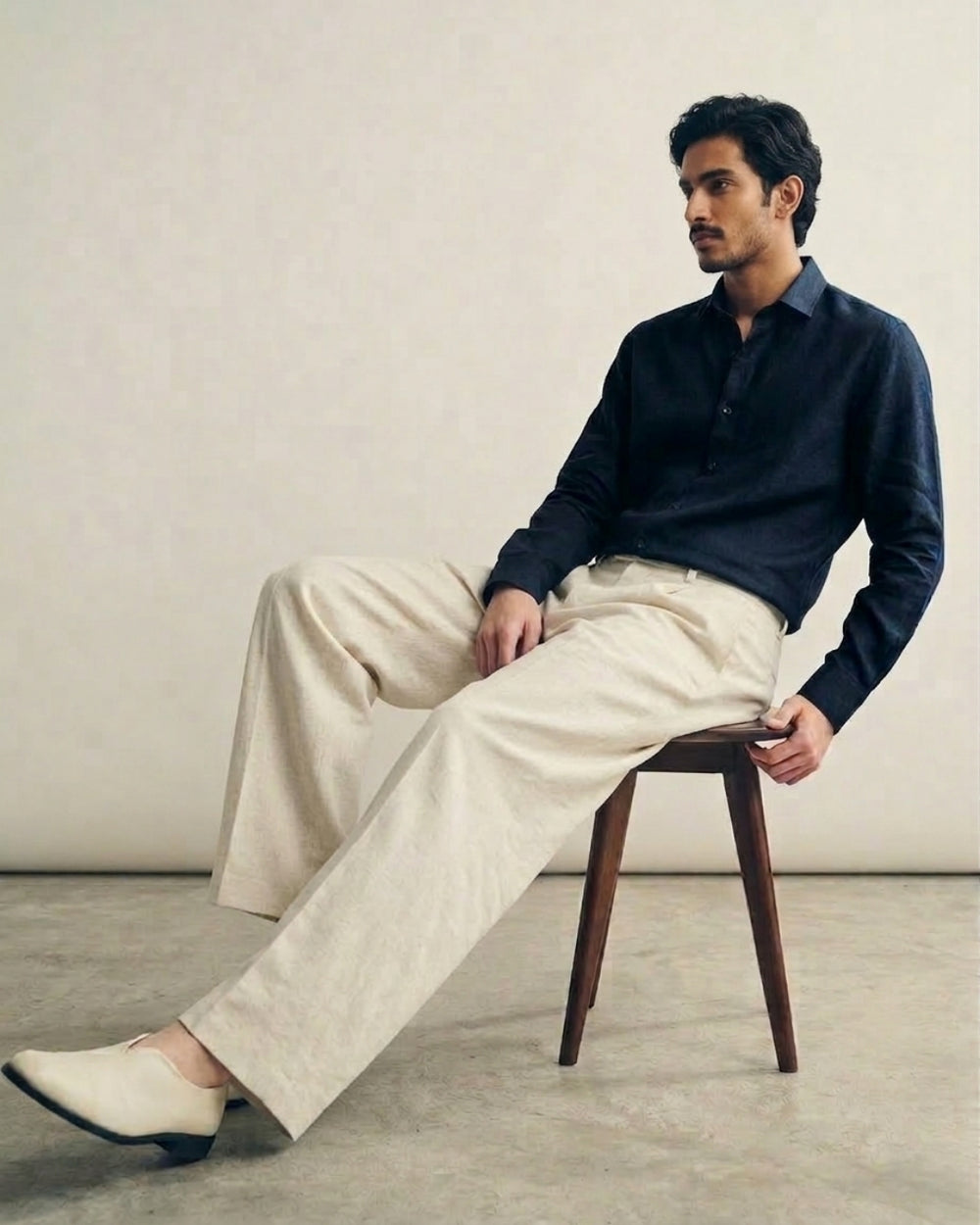 Man seated on a wooden chair in the Aakar Blue navy pure linen shirt by Tyra with cream trousers