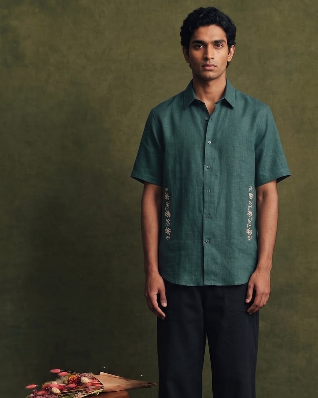Deep teal green linen short-sleeve shirt with a classic collar, button-front closure, and intricate cream embroidered detailing along both sides of the front, paired with black trousers.