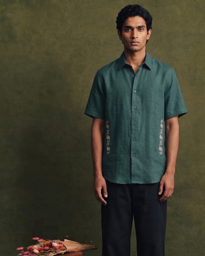 Deep teal green linen short-sleeve shirt with a classic collar, button-front closure, and intricate cream embroidered detailing along both sides of the front, paired with black trousers.