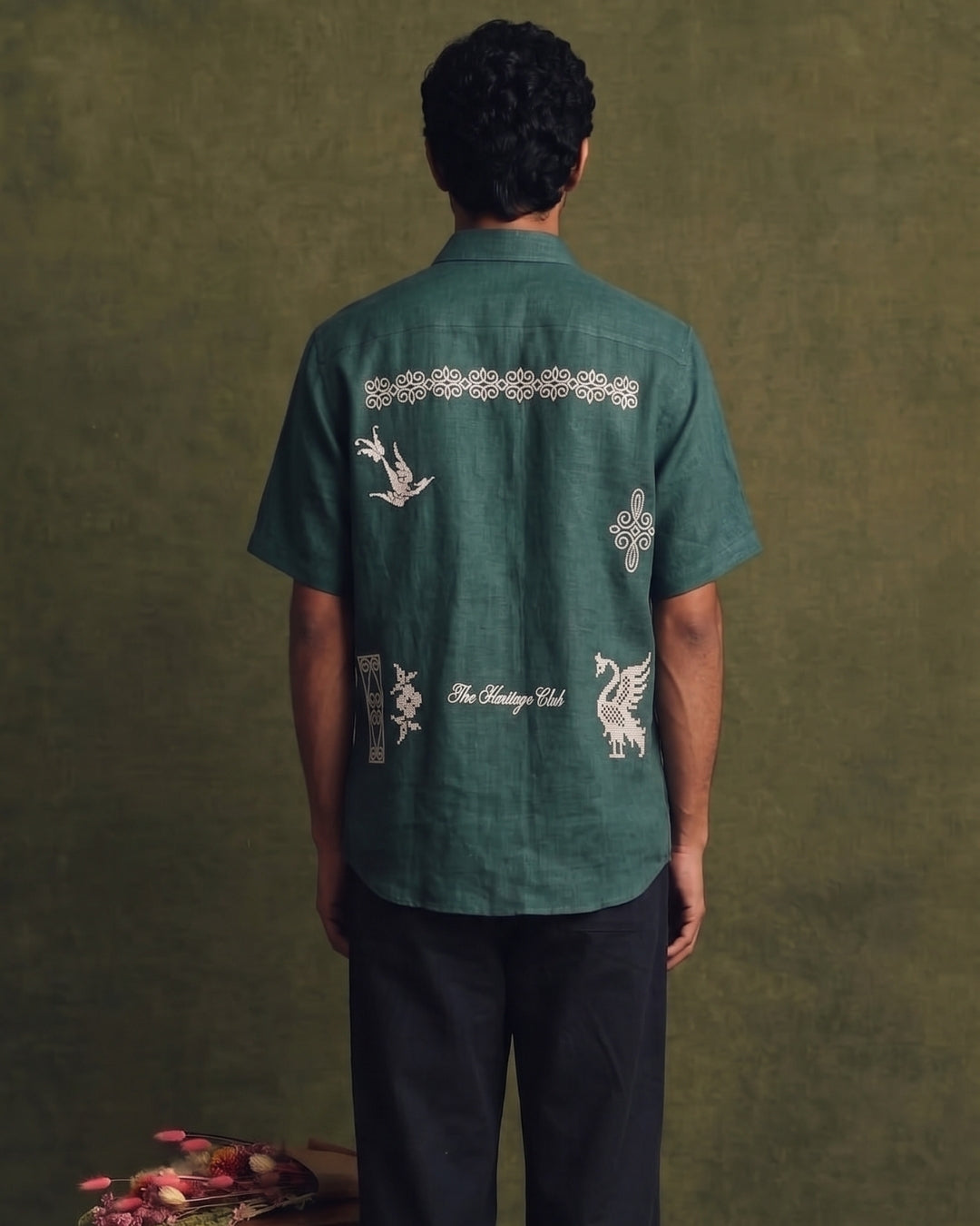 Deep teal green linen short-sleeve shirt with a classic collar and button-front closure, featuring intricate cream embroidered motifs and decorative artwork across the back, paired with black trousers.