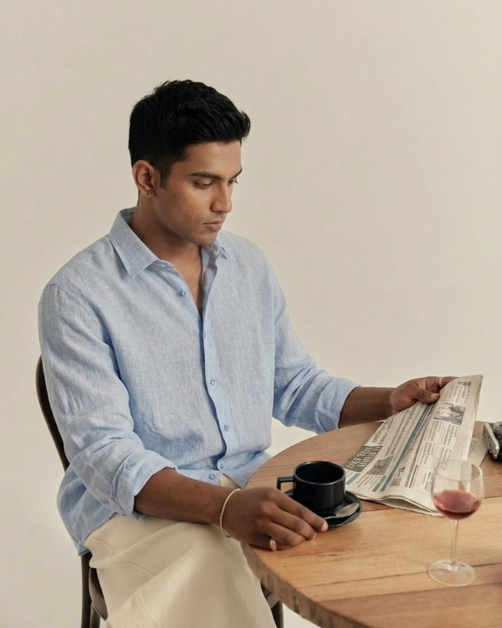 Man seated at a wooden table with a newspaper and coffee, wearing the First Light Blue pure linen shirt by Tyra