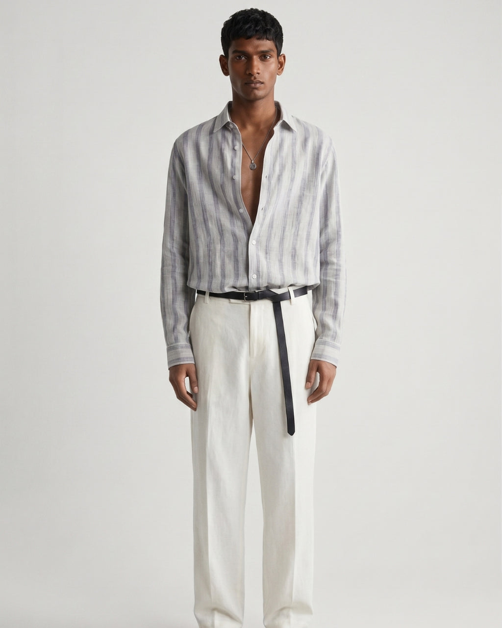 Full-length front view of a male model wearing a mist-grey striped linen shirt with a relaxed fit, open collar, and button-front closure, paired with off-white tailored trousers, a black leather belt, and black leather sandals against a minimalist light neutral studio background.