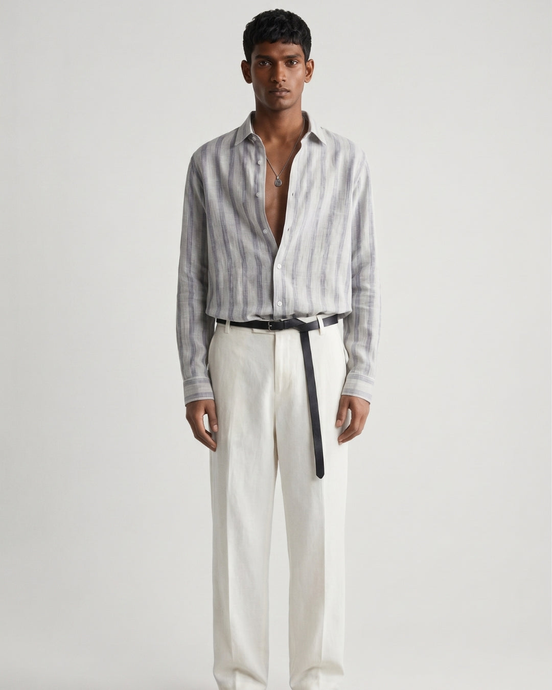 Full-length front view of a male model wearing a mist-grey striped linen shirt with a relaxed fit, open collar, and button-front closure, paired with off-white tailored trousers, a black leather belt, and black leather sandals against a minimalist light neutral studio background.