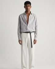 Full-length front view of a male model wearing a mist-grey striped linen shirt with a relaxed fit, open collar, and button-front closure, paired with off-white tailored trousers, a black leather belt, and black leather sandals against a minimalist light neutral studio background.