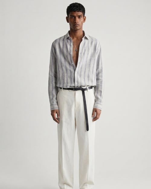 Full-length front view of a male model wearing a mist-grey striped linen shirt with a relaxed fit, open collar, and button-front closure, paired with off-white tailored trousers, a black leather belt, and black leather sandals against a minimalist light neutral studio background.