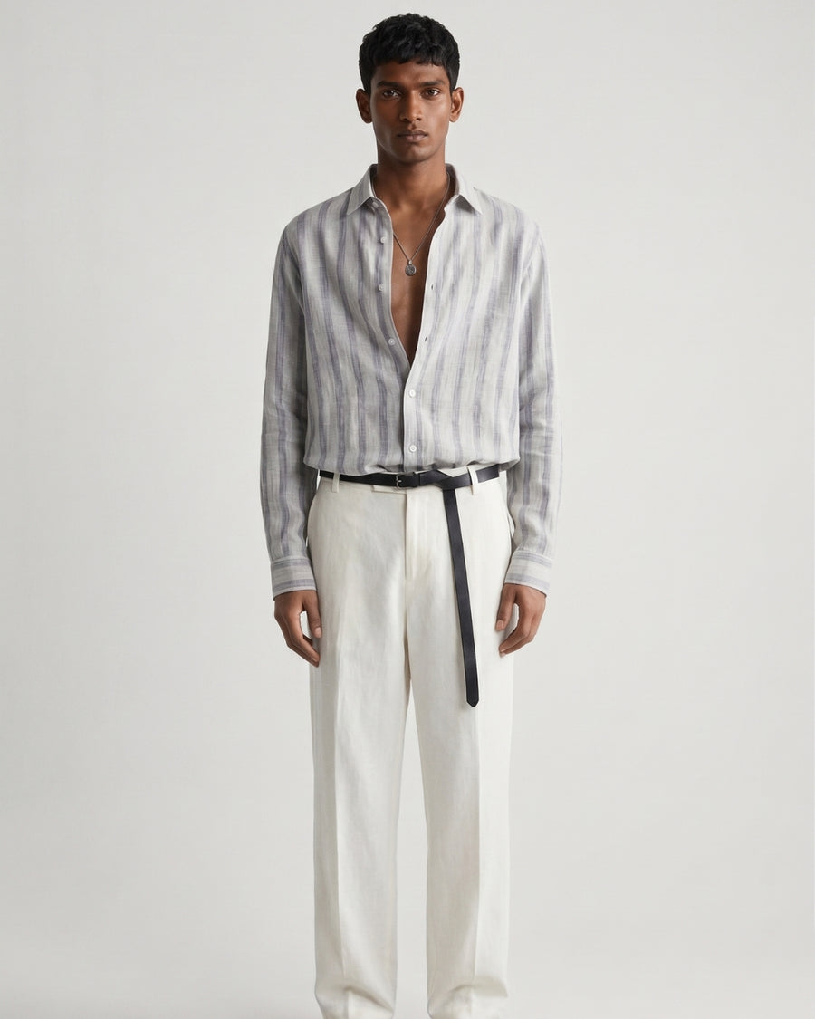 Full-length front view of a male model wearing a mist-grey striped linen shirt with a relaxed fit, open collar, and button-front closure, paired with off-white tailored trousers, a black leather belt, and black leather sandals against a minimalist light neutral studio background.