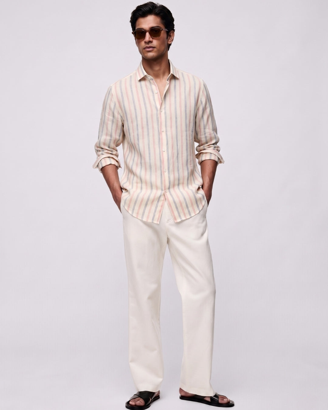 Full-length front view of a male model wearing a rosé striped linen shirt featuring blush pink and muted blue vertical stripes, with a classic collar and button-front closure, paired with off-white tailored trousers, black leather sandals, and brown sunglasses against a clean light-grey studio background.