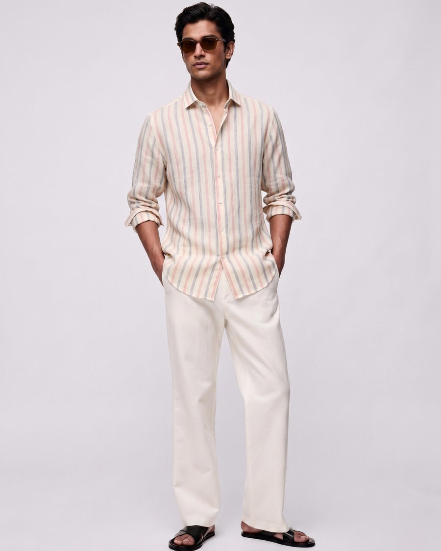 Full-length front view of a male model wearing a rosé striped linen shirt featuring blush pink and muted blue vertical stripes, with a classic collar and button-front closure, paired with off-white tailored trousers, black leather sandals, and brown sunglasses against a clean light-grey studio background.