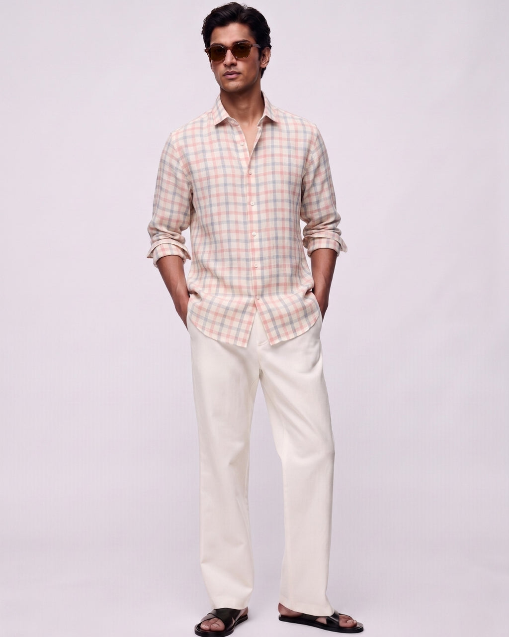 Full-length front view of a male model wearing a rosé grid linen shirt featuring soft pink and muted blue check patterns, with a classic collar and button-front closure, paired with off-white tailored trousers, black leather sandals, and brown sunglasses against a clean light-grey studio background.