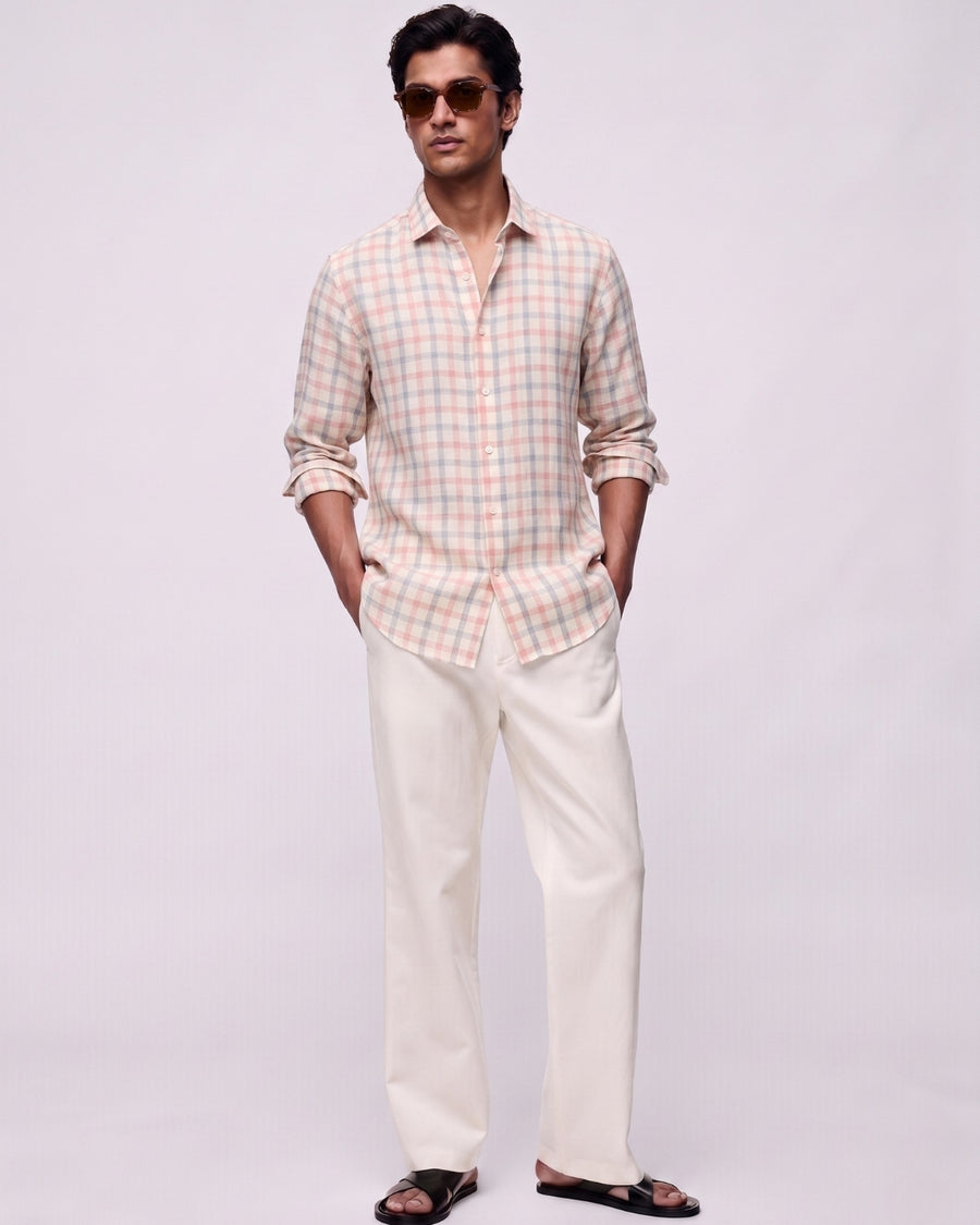 Full-length front view of a male model wearing a rosé grid linen shirt featuring soft pink and muted blue check patterns, with a classic collar and button-front closure, paired with off-white tailored trousers, black leather sandals, and brown sunglasses against a clean light-grey studio background.