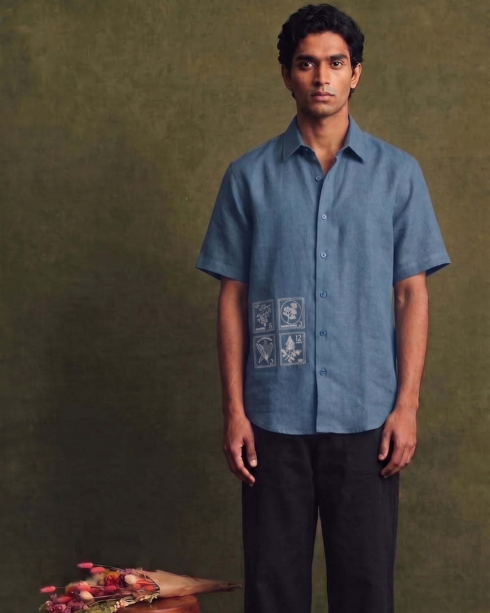 Dusty blue linen short-sleeve shirt with a classic collar, button-front closure, and four-panel botanical embroidery detail on the lower front, paired with black trousers.