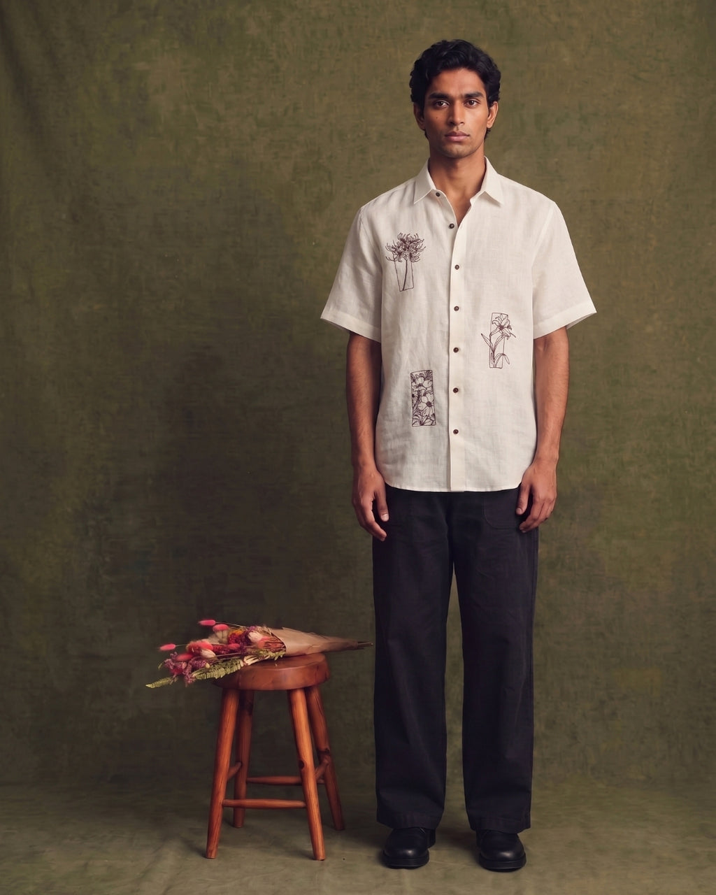 Alt text: Off-white linen short-sleeve shirt with a classic collar, button-front closure, and hand-drawn floral embroidery details, paired with black trousers.