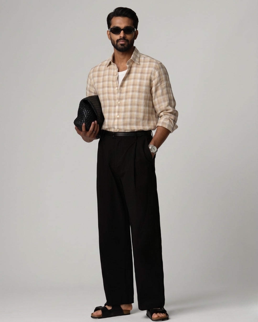 Full-length front view of a male model wearing a Kora Brown Grid Linen Shirt in a soft beige-brown check pattern, paired with black tailored trousers, black sandals, and dark sunglasses, holding a woven black clutch against a light grey studio background.