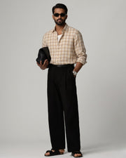 Full-length front view of a male model wearing a Kora Brown Grid Linen Shirt in a soft beige-brown check pattern, paired with black tailored trousers, black sandals, and dark sunglasses, holding a woven black clutch against a light grey studio background.