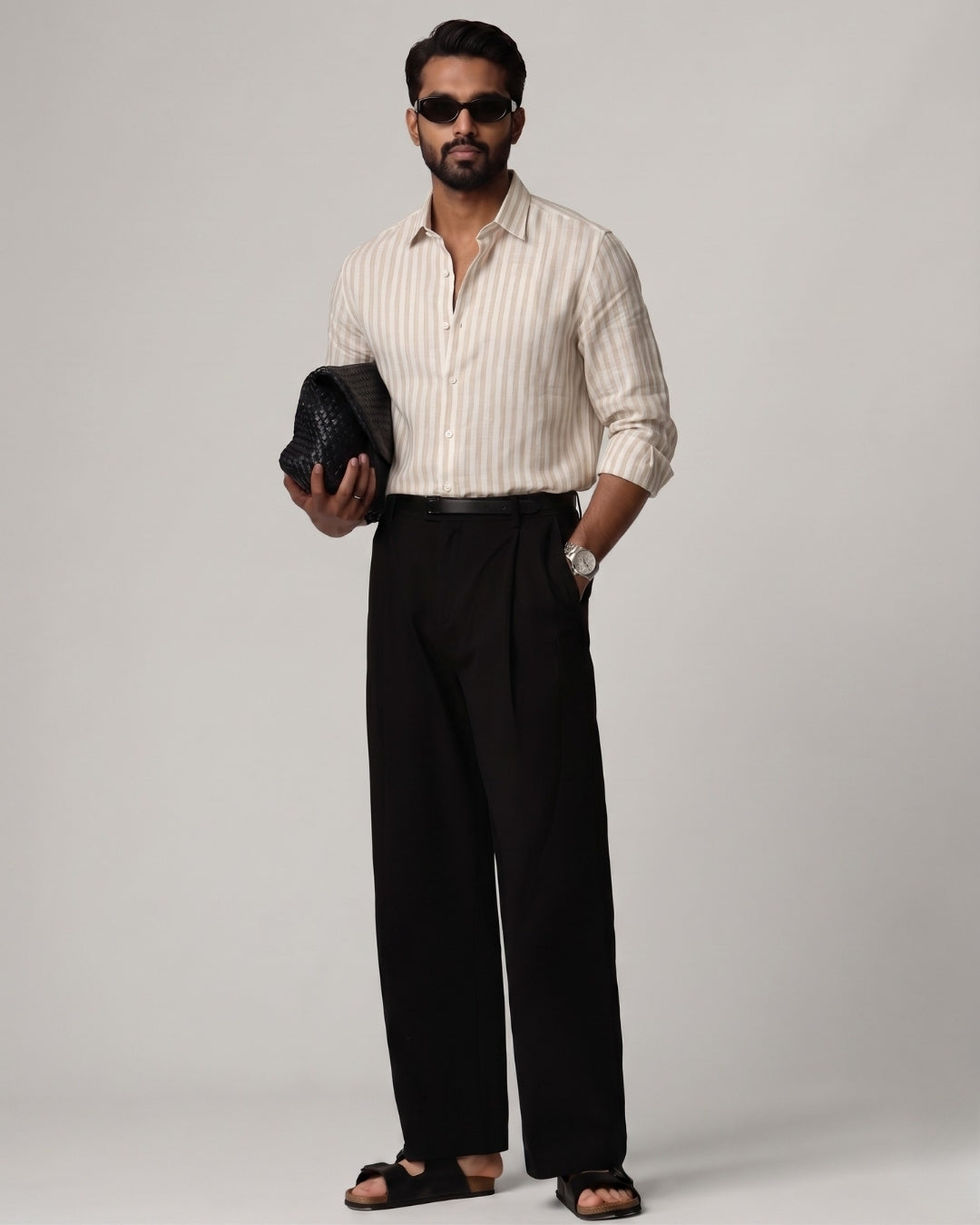 Full-length front view of a male model wearing a Brown Trace Striped Linen Shirt with subtle beige and brown vertical stripes, paired with black tailored trousers, black sandals, dark sunglasses, and a woven black clutch against a light grey studio background.