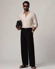 Full-length front view of a male model wearing a Brown Trace Striped Linen Shirt with subtle beige and brown vertical stripes, paired with black tailored trousers, black sandals, dark sunglasses, and a woven black clutch against a light grey studio background.