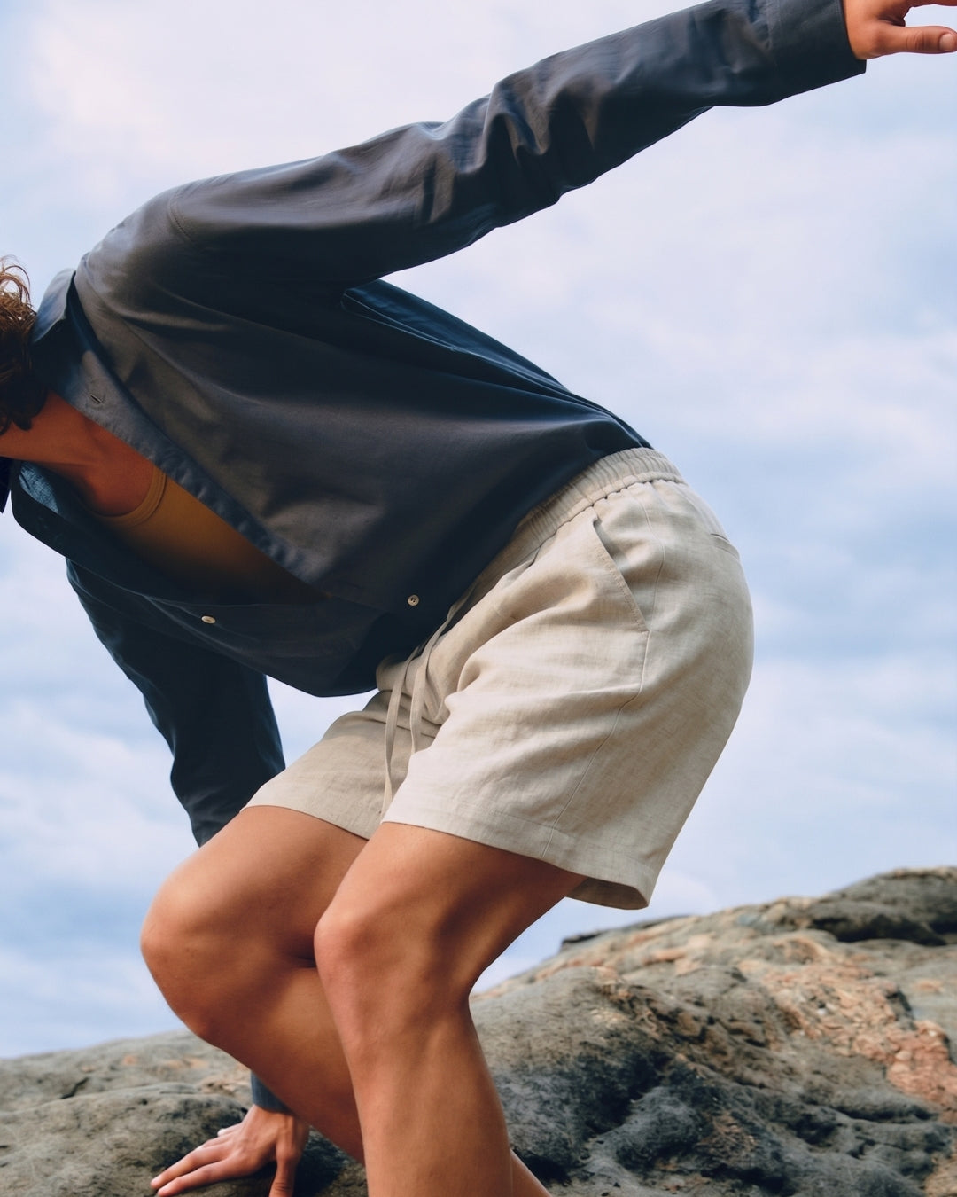 Model wearing Natural Casual Shorts in lightweight natural beige linen with an elasticated drawstring waistband and relaxed fit, styled with a dark overshirt while posed on coastal rocks under soft daylight.