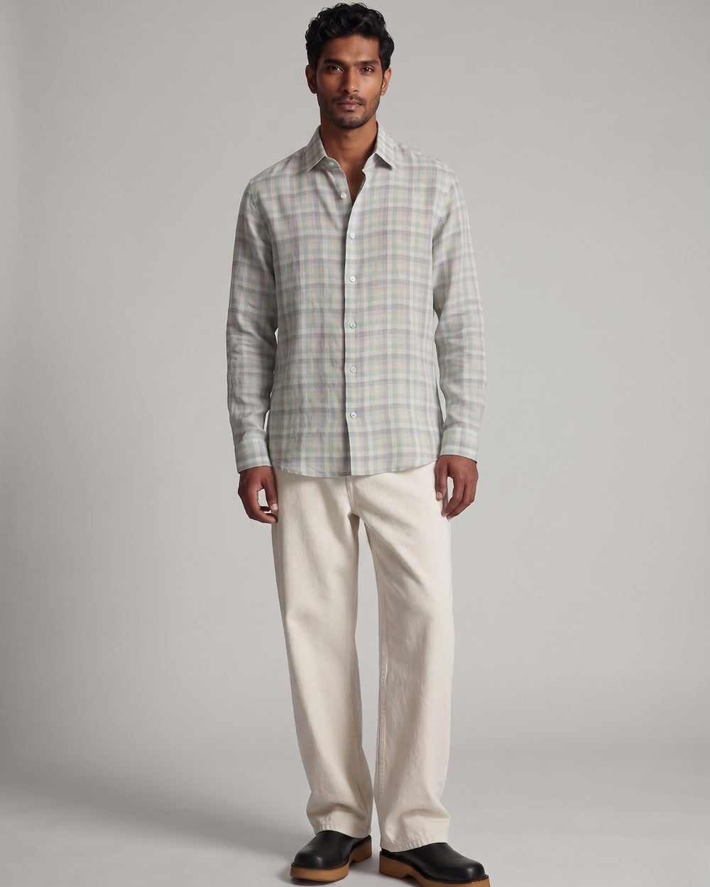 Full-length front view of a male model wearing the Still Grid Linen Shirt featuring a soft sage, stone, and ivory grid pattern, paired with off-white relaxed-fit trousers and black leather boots against a light grey studio background.