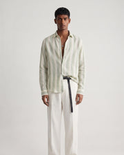 Full-length front view of a male model wearing a sage green striped linen shirt with a relaxed fit, classic collar, and button-front closure, paired with off-white tailored trousers, a black leather belt, and black leather sandals against a minimalist light neutral studio background.