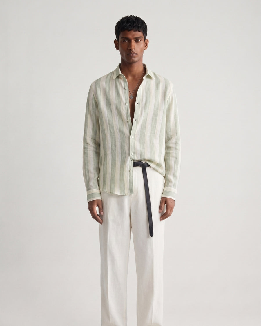 Full-length front view of a male model wearing a sage green striped linen shirt with a relaxed fit, classic collar, and button-front closure, paired with off-white tailored trousers, a black leather belt, and black leather sandals against a minimalist light neutral studio background.