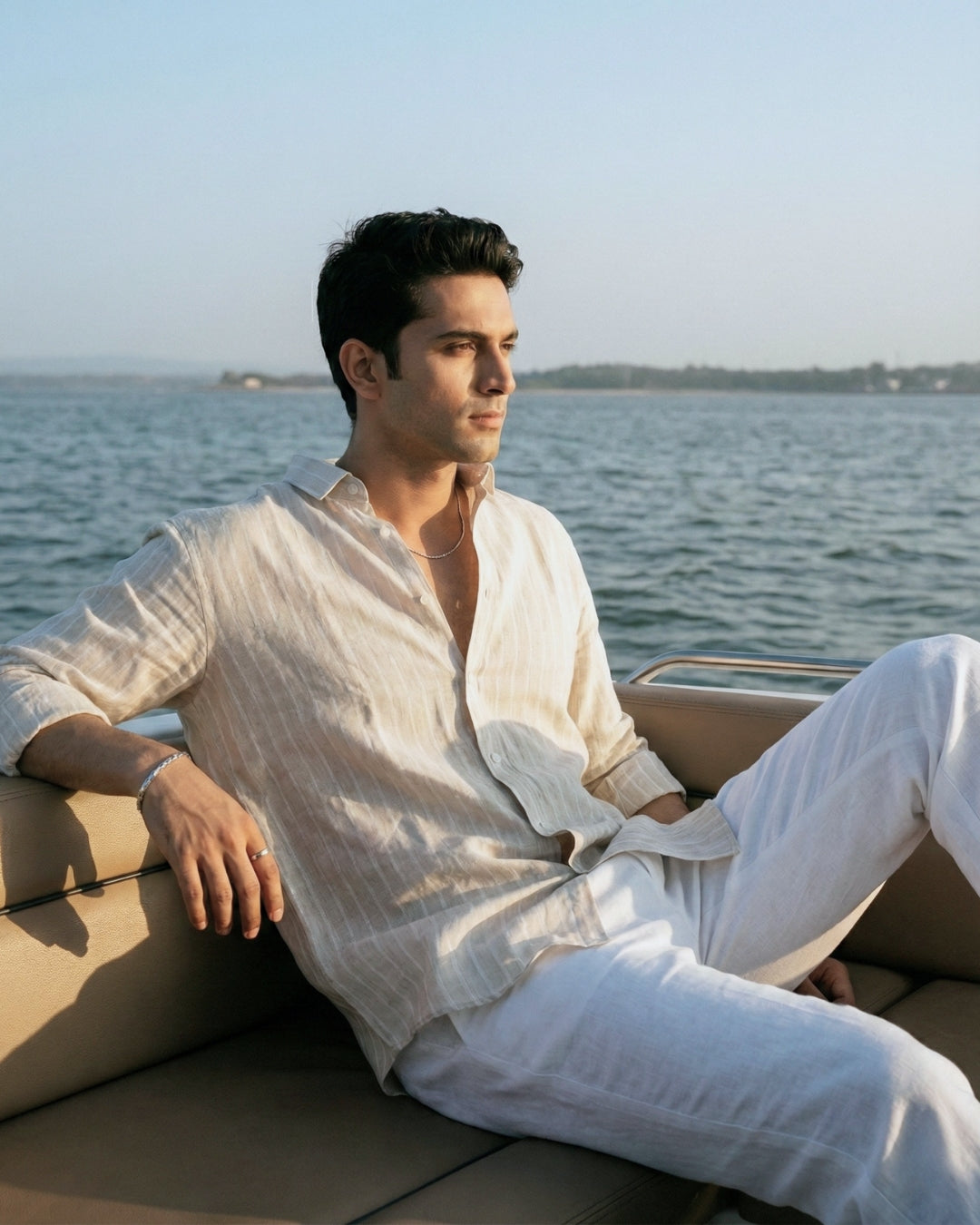 Model wearing a Tyra lightweight striped linen shirt and relaxed white linen trousers, styled in a coastal setting with soft natural light, showcasing breathable luxury summer menswear.