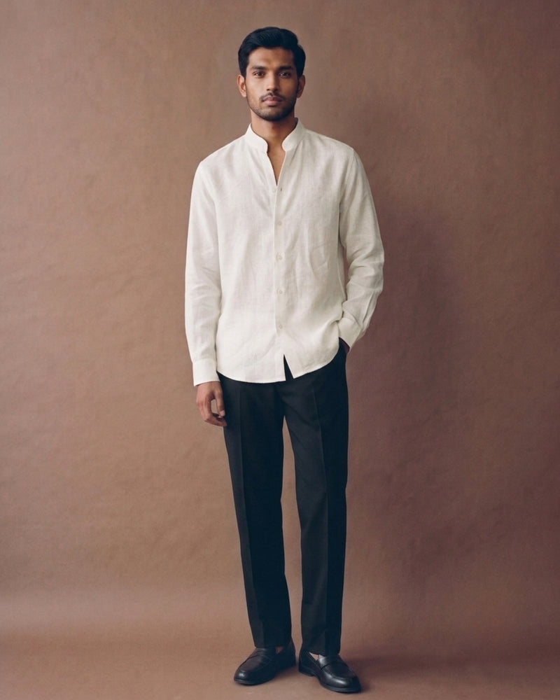 Male model wearing an ivory linen shirt with a mandarin collar, button-down front, and curved hem, paired with black trousers against a warm brown studio backdrop.