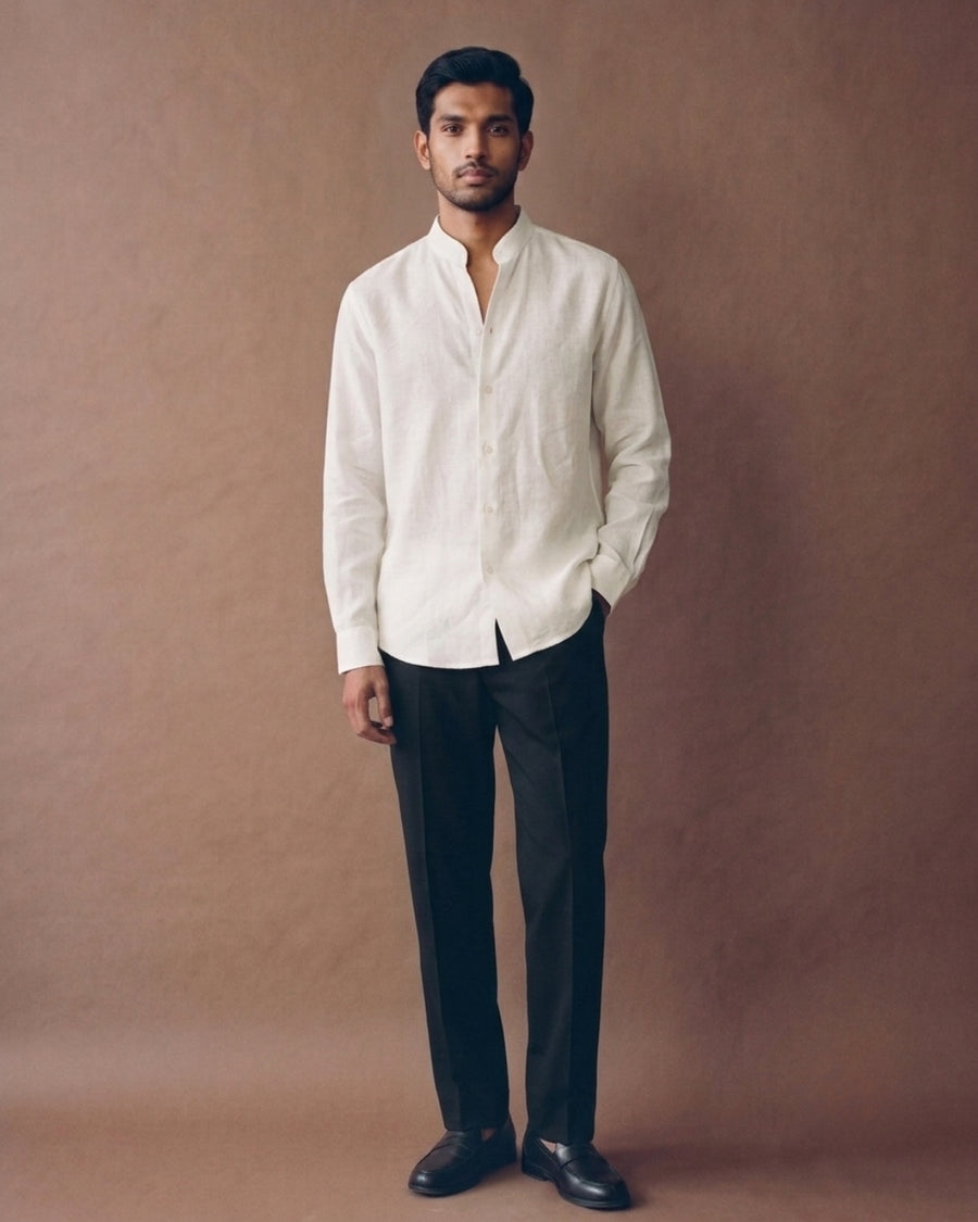 Male model wearing an ivory linen shirt with a mandarin collar, button-down front, and curved hem, paired with black trousers against a warm brown studio backdrop.
