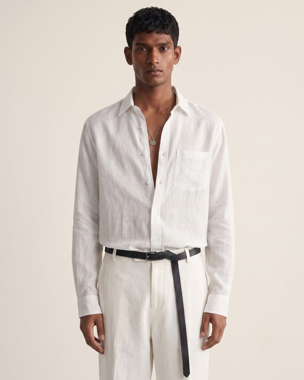 A full-length portrait of a man standing against a soft, warm beige backdrop. He wears a relaxed white linen button-down shirt with the top buttons undone, paired with high-waisted white trousers and a slim black leather belt with a long hanging strap. A simple silver pendant necklace rests on his chest. His expression is neutral, with his arms relaxed at his sides. The image features soft, diffused lighting and a clean, minimalist aesthetic that emphasizes the natural texture of the linen fa