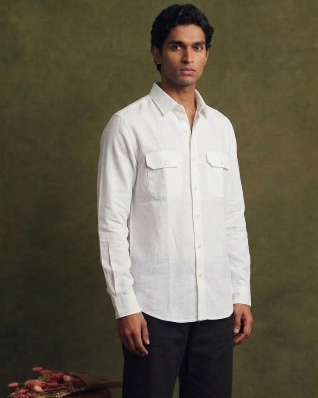  White linen shirt with long sleeves, pointed collar, button-front closure, and two flap chest pockets, worn by a male model with black trousers.