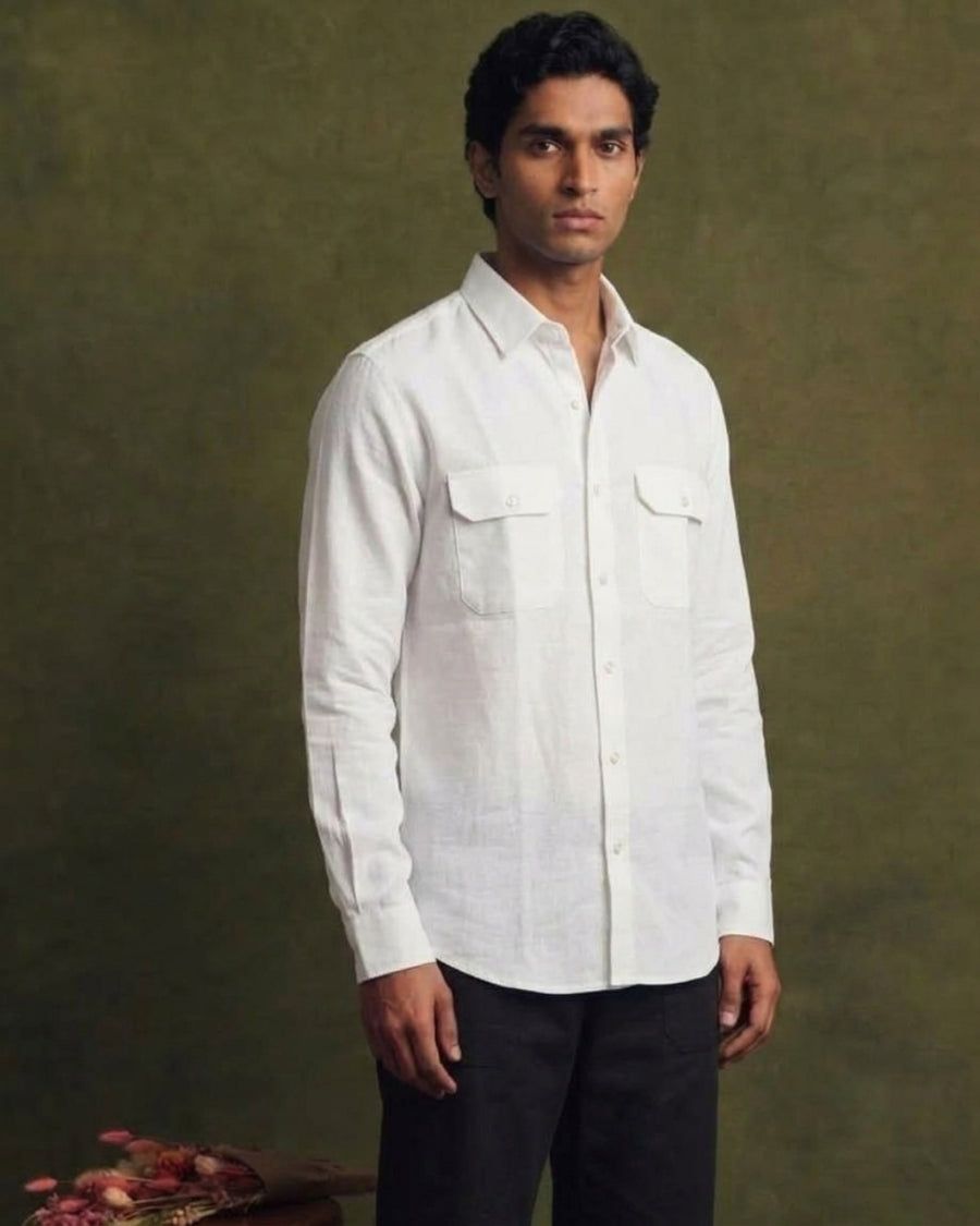  White linen shirt with long sleeves, pointed collar, button-front closure, and two flap chest pockets, worn by a male model with black trousers.