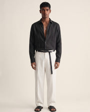 Man in the Midnight Black pure linen shirt by Tyra, worn open with white trousers and a black leather belt against a warm beige backdrop