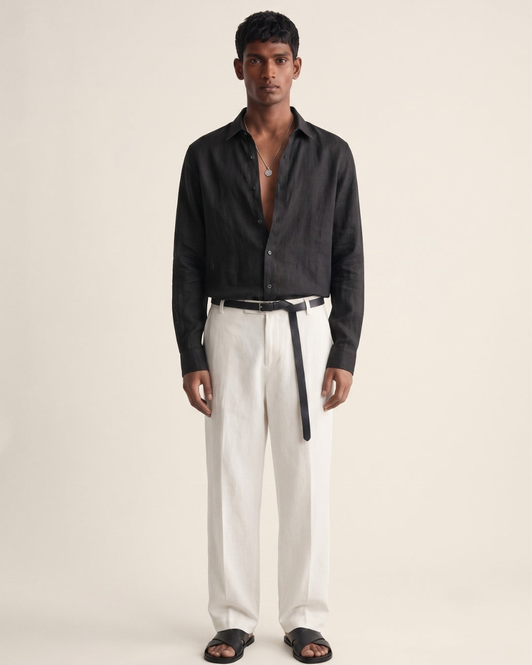A full-length portrait of a man standing against a soft, warm beige backdrop. He wears a relaxed black linen button-down shirt with the top buttons undone, paired with high-waisted white trousers, a slim black leather belt with a long hanging strap, and black leather slide sandals. A simple silver pendant necklace adds a subtle accent. He stands facing forward with a neutral expression and his arms relaxed at his sides. Soft, diffused lighting and a minimalist setting highlight the contrast b