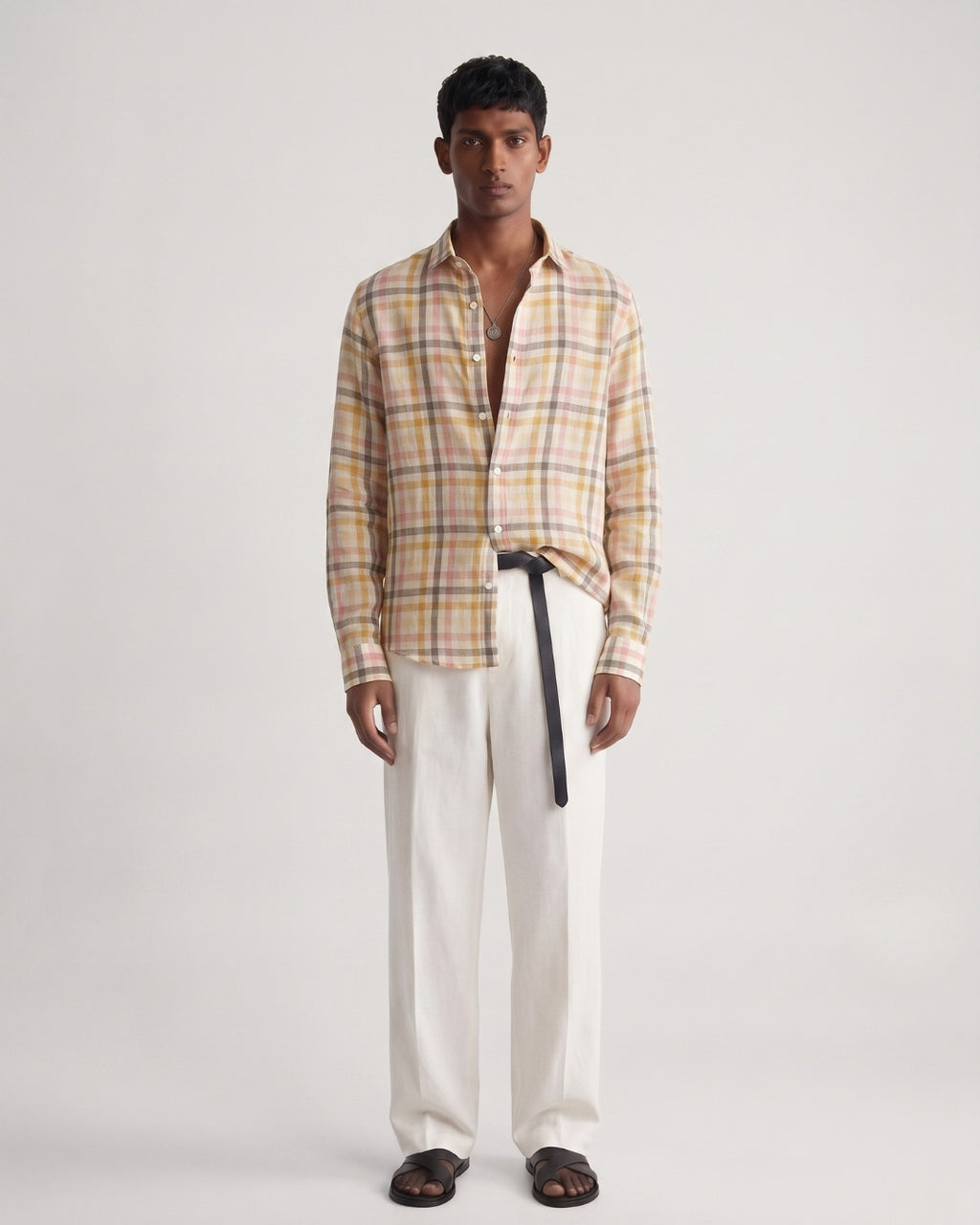 Full-length front view of a male model wearing a pastel plaid linen shirt featuring sand, blush pink, and muted yellow checks, with a classic collar and button-front closure, paired with off-white tailored trousers, a black leather belt, and black leather sandals against a minimalist light neutral studio background.