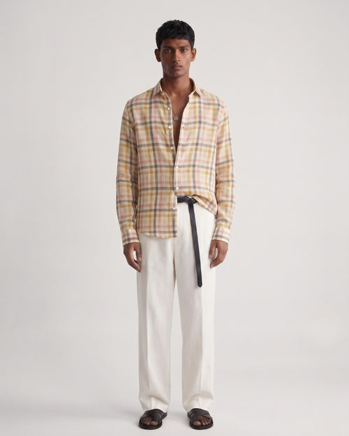 Full-length front view of a male model wearing a pastel plaid linen shirt featuring sand, blush pink, and muted yellow checks, with a classic collar and button-front closure, paired with off-white tailored trousers, a black leather belt, and black leather sandals against a minimalist light neutral studio background.