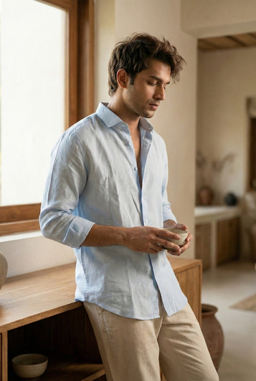 Man holding a coffee mug by a window, wearing the Asmaan sky blue pure linen shirt by Tyra