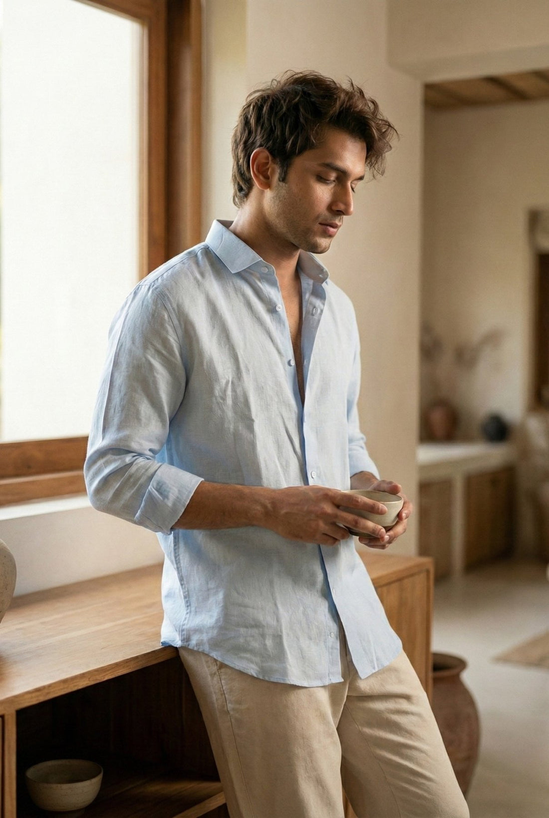 Man holding a coffee mug by a window, wearing the Asmaan sky blue pure linen shirt by Tyra