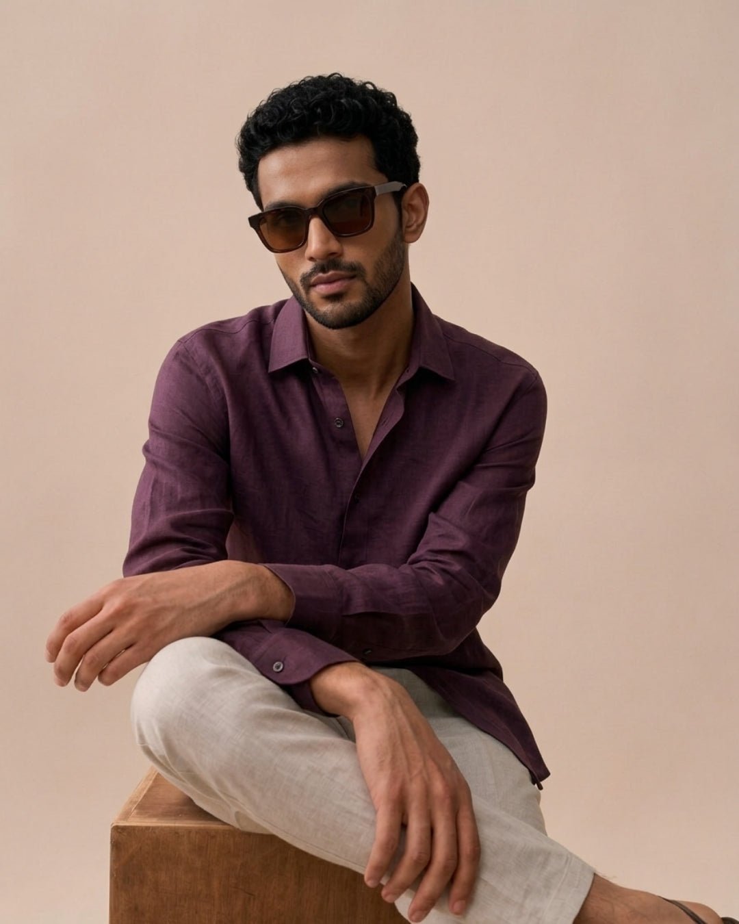Man seated on a wooden block against a neutral backdrop, wearing a deep maroon long-sleeve shirt with light beige trousers and sunglasses; posed casually with a relaxed, refined aesthetic.
Tyra