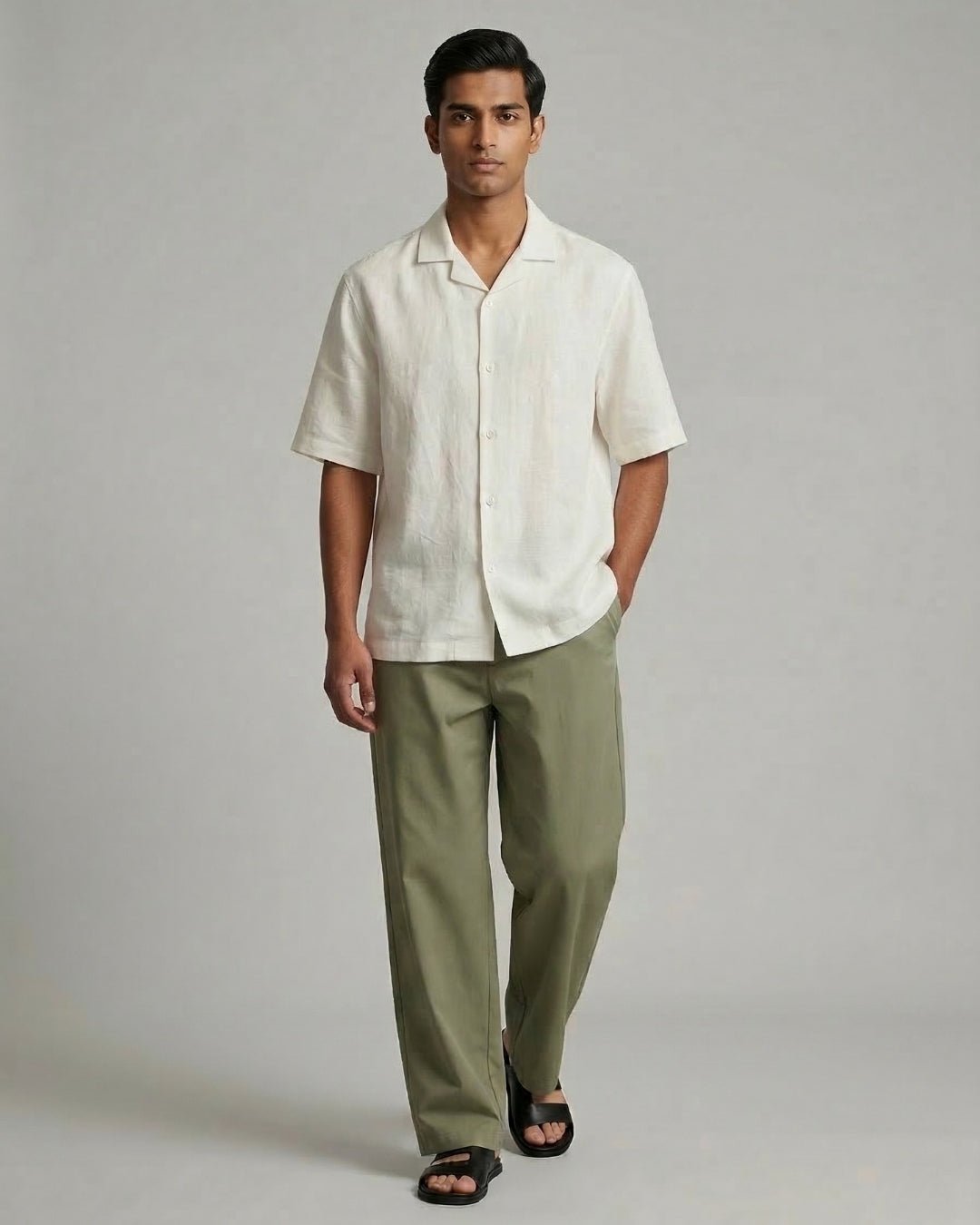 Man walking against a neutral grey backdrop, wearing an off-white short-sleeve button-up shirt paired with olive green trousers and black sandals; styled in a clean, minimal aesthetic.
Tyra