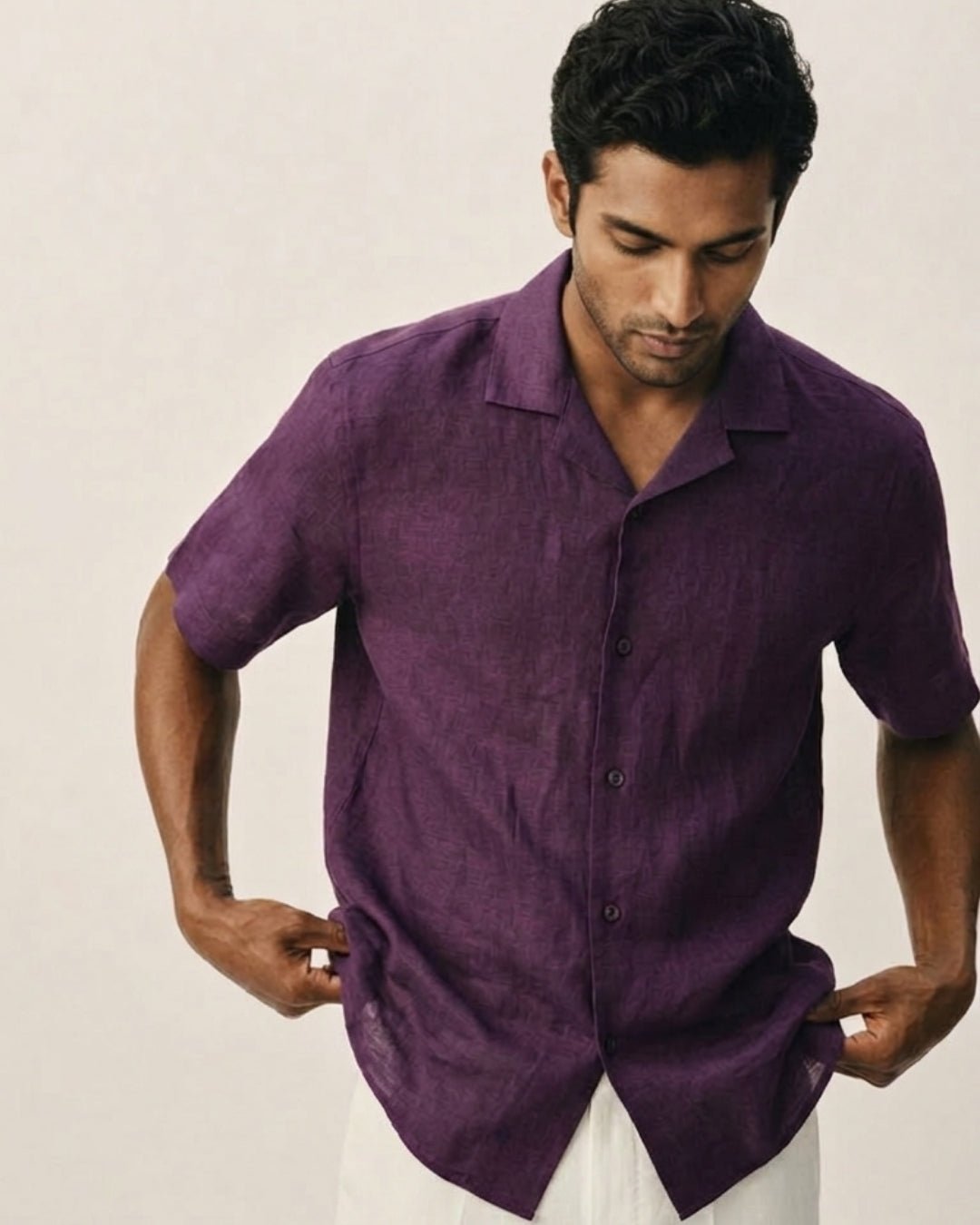 Man wearing a deep purple short-sleeve button-up shirt with a subtle textured pattern, adjusting the hem; paired with light-colored trousers against a neutral background, styled in a minimal, refined aesthetic.
Tyra