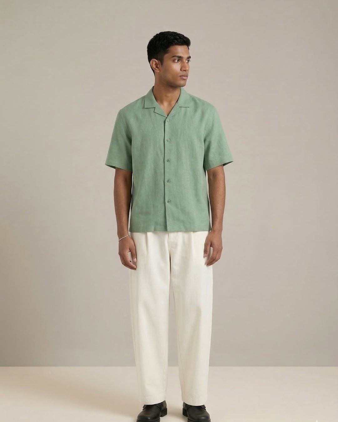 Man standing against a neutral backdrop, wearing a sage green short-sleeve button-up shirt paired with off-white relaxed trousers and black shoes; styled in a clean, minimal aesthetic.
Tyra