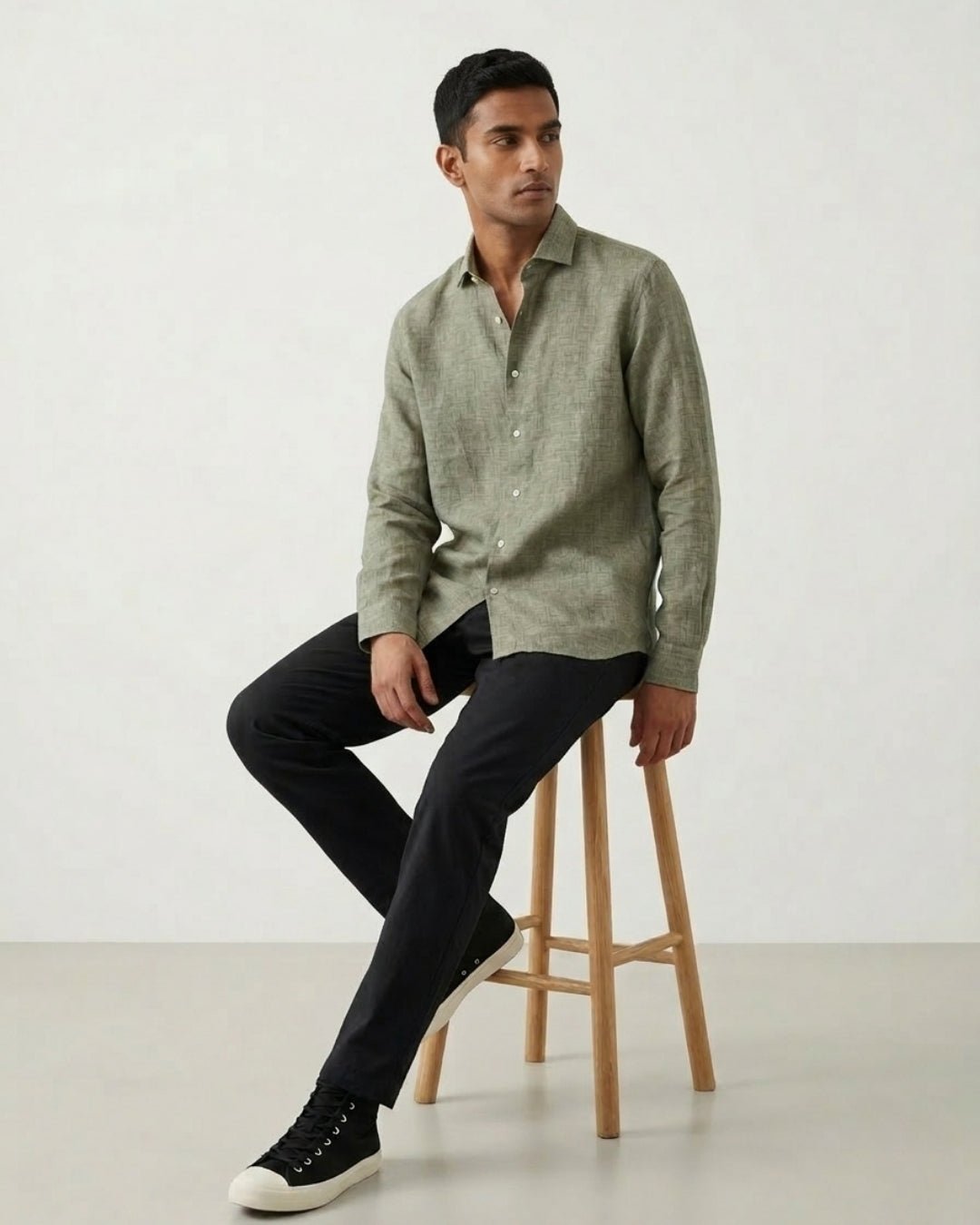 Man seated on a wooden stool against a neutral backdrop, wearing a textured sage green long-sleeve shirt paired with black trousers and black sneakers; styled in a clean, minimal aesthetic.
Tyra