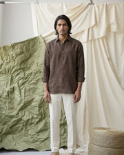 Man in the PopOver Brown long-sleeve linen shirt by Tyra with white trousers against a layered fabric backdrop