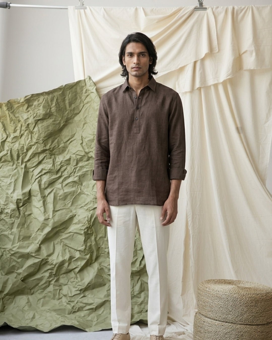 Man standing against layered fabric backdrops in neutral tones, wearing a brown long-sleeve band-collar shirt with a button placket, paired with off-white tailored trousers and beige shoes; styled in a minimal, earthy studio setting.
Tyra