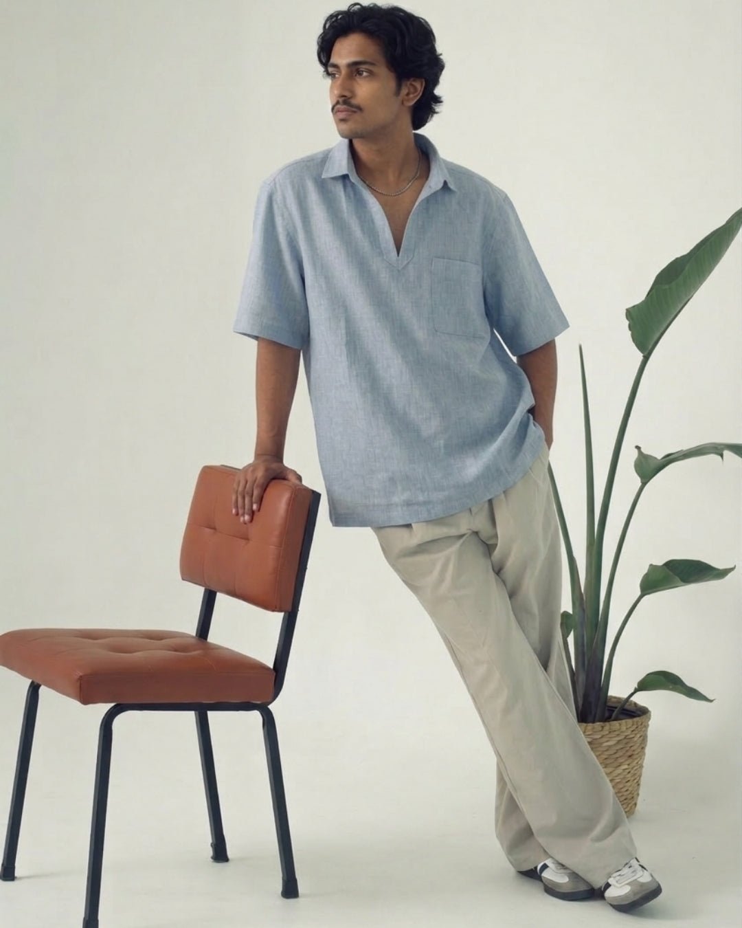Man leaning against a brown chair, wearing a light blue short-sleeve shirt with an open collar and chest pocket, paired with relaxed beige trousers and casual sneakers; styled in a clean, minimal indoor setting with a potted plant.
Tyra