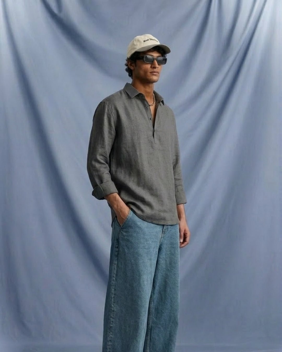 Man standing against a soft blue draped backdrop, wearing a charcoal grey long-sleeve shirt with an open collar, paired with relaxed blue denim jeans; styled with a beige cap, sunglasses, and a minimal, contemporary aesthetic.
Tyra