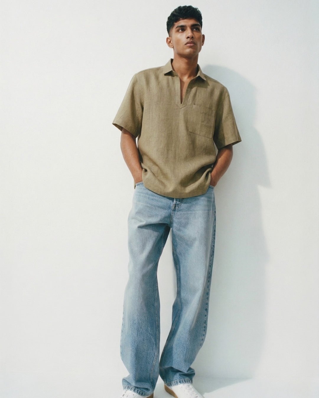 Man leaning against a light wall wearing an olive short-sleeve shirt with an open collar and chest pocket, paired with relaxed light-wash jeans and white sneakers; hands in pockets, styled in a casual, minimal aesthetic. 
Tyra
