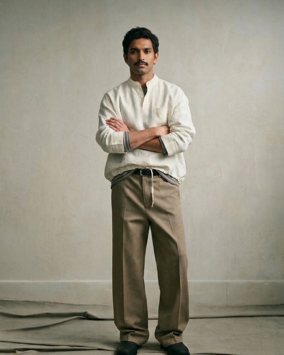 Man standing against a neutral beige backdrop, wearing a relaxed off-white band-collar shirt with rolled sleeves, olive-brown pleated trousers, and black shoes; arms crossed, styled in a minimal, earthy aesthetic.
Tyra