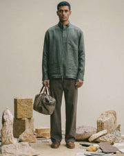 Man holding a duffel bag in the Rove Olive band-collar pure linen shirt by Tyra with brown trousers, set among stone blocks