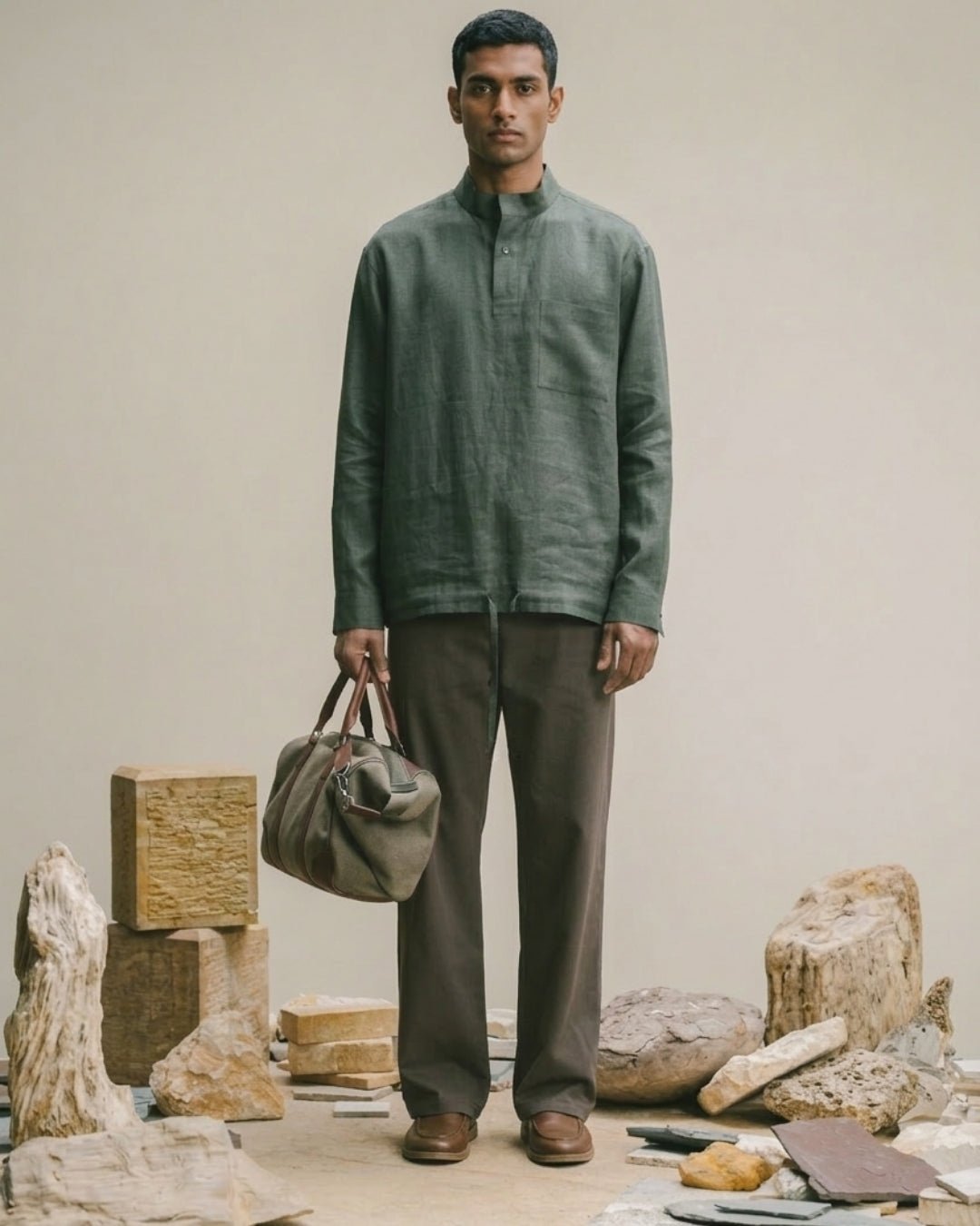 Man standing against a neutral backdrop wearing an earthy green band-collar kurta-style shirt with matching tailored trousers and brown shoes, holding a structured duffel bag; surrounded by natural stone and wood elements, evoking a raw, minimalist aesthetic.
Tyra