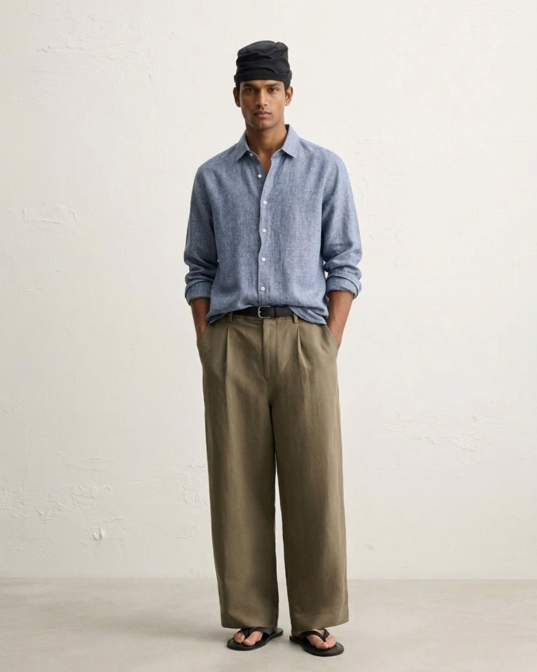 Man standing against a textured neutral wall, wearing a light blue long-sleeve button-up shirt tucked into olive wide-leg trousers with a belt, paired with black sandals and a black cap; styled in a relaxed, minimal aesthetic.
Tyra